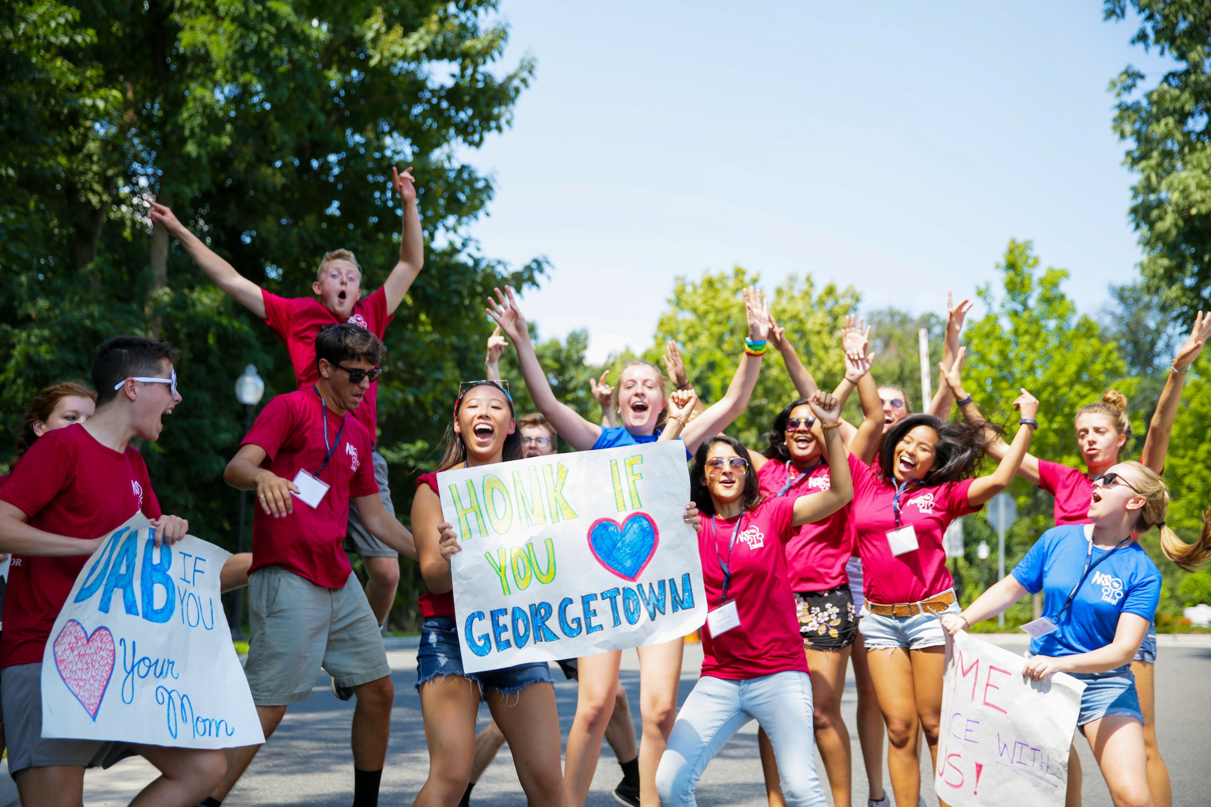 A group of Orientation leaders welcome new students to campus. They are jumping in the air, and one holds a sign that says 