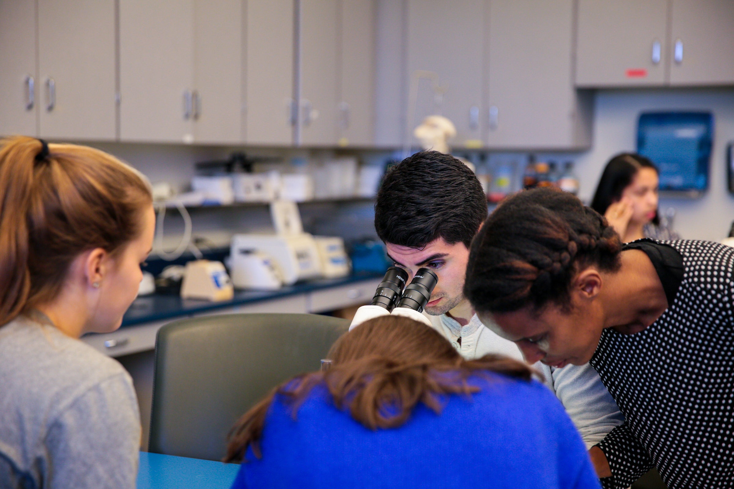 Four students work around a table, while one of them looks into a microscope.
