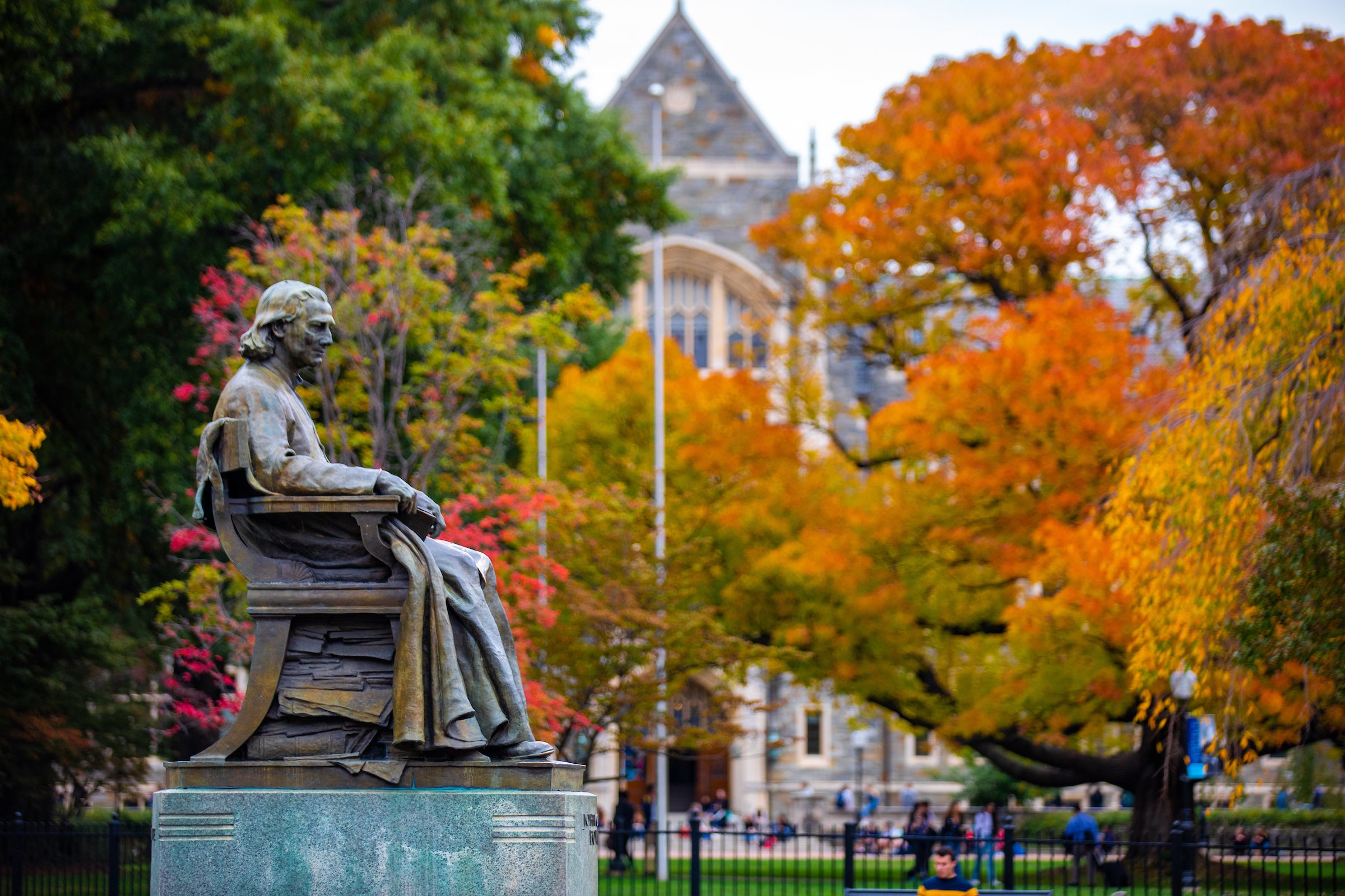 A statue of John Carroll is in the foreground, with White-Gravenor Hall and trees with yellow and orange leaves in the background.
