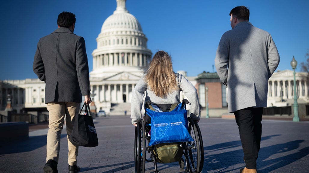 Three students, including one using a wheelchair, walk and wheel in front of the US Capitol building.