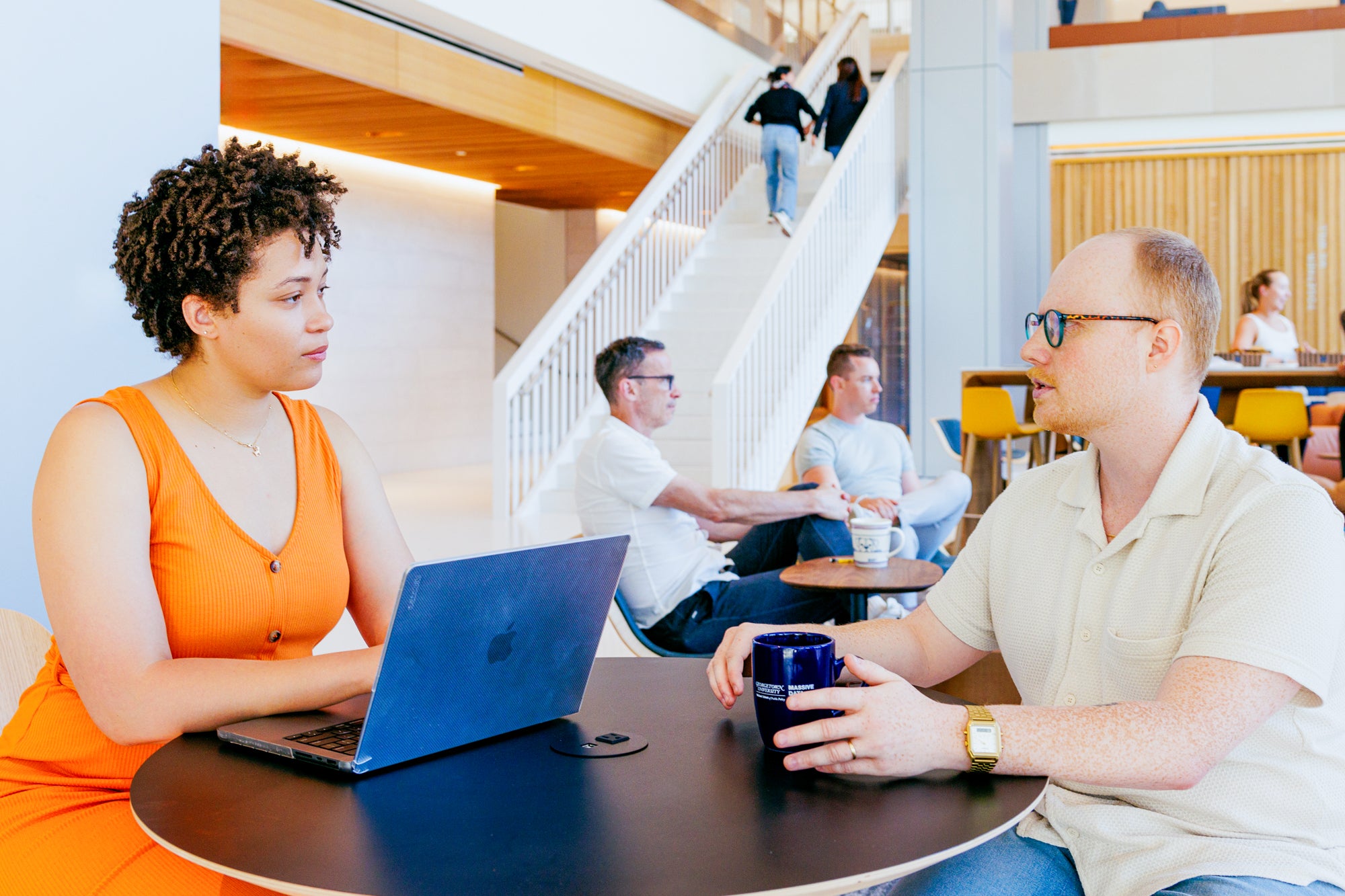 Two students speak to each other at a table in the lobby of the McCourt School building. One student has a laptop, and the other has a mug.