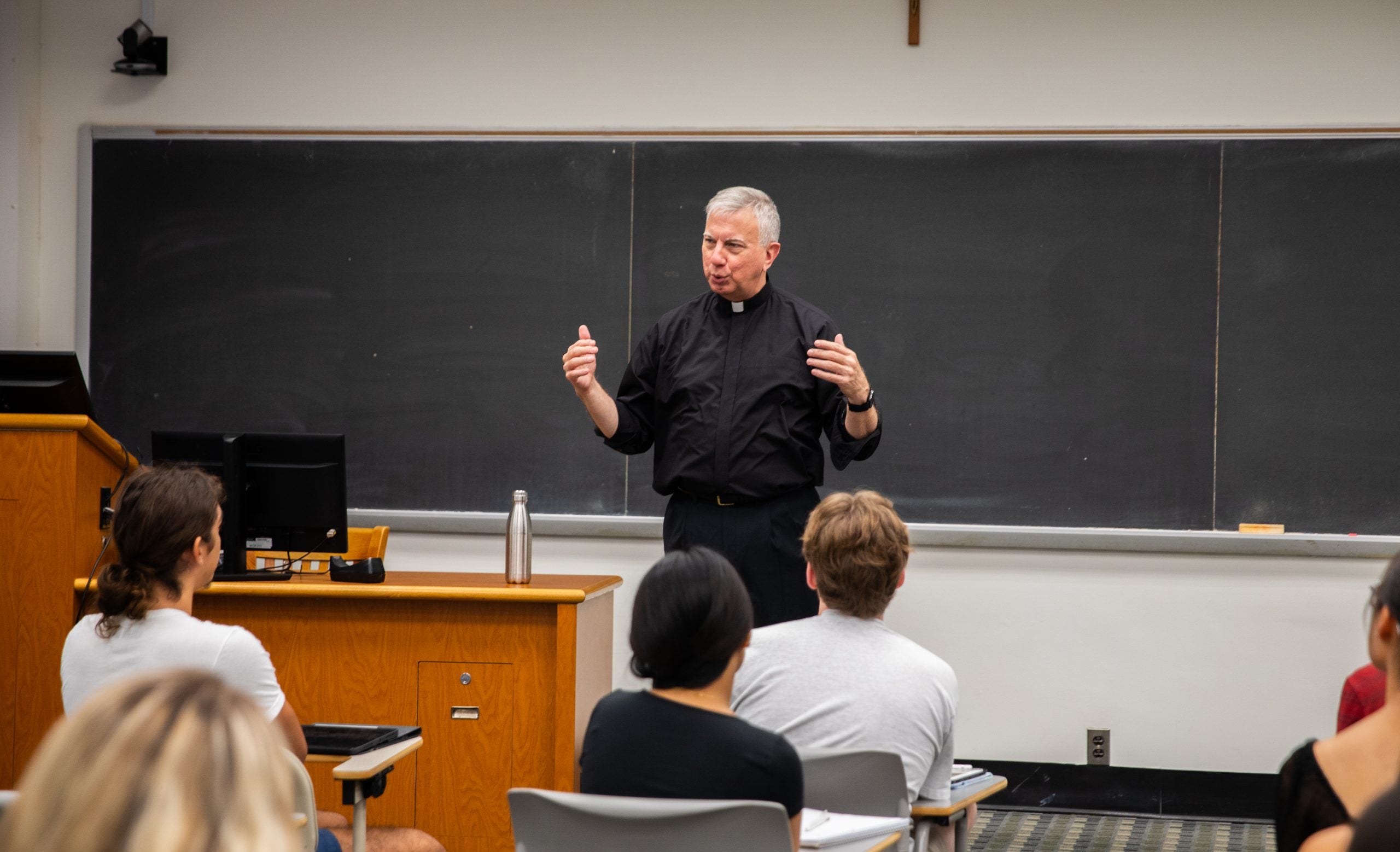 A Jesuit Professor stands at the front of a classroom while teaching.