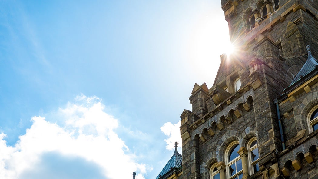 A zoomed-in image of the Healy Hall clock tower with sun shining behind it.