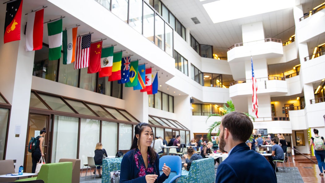 Two students talk to each other in the Intercultural Center Galleria. Behind them, the atrium displays flags from many nations.