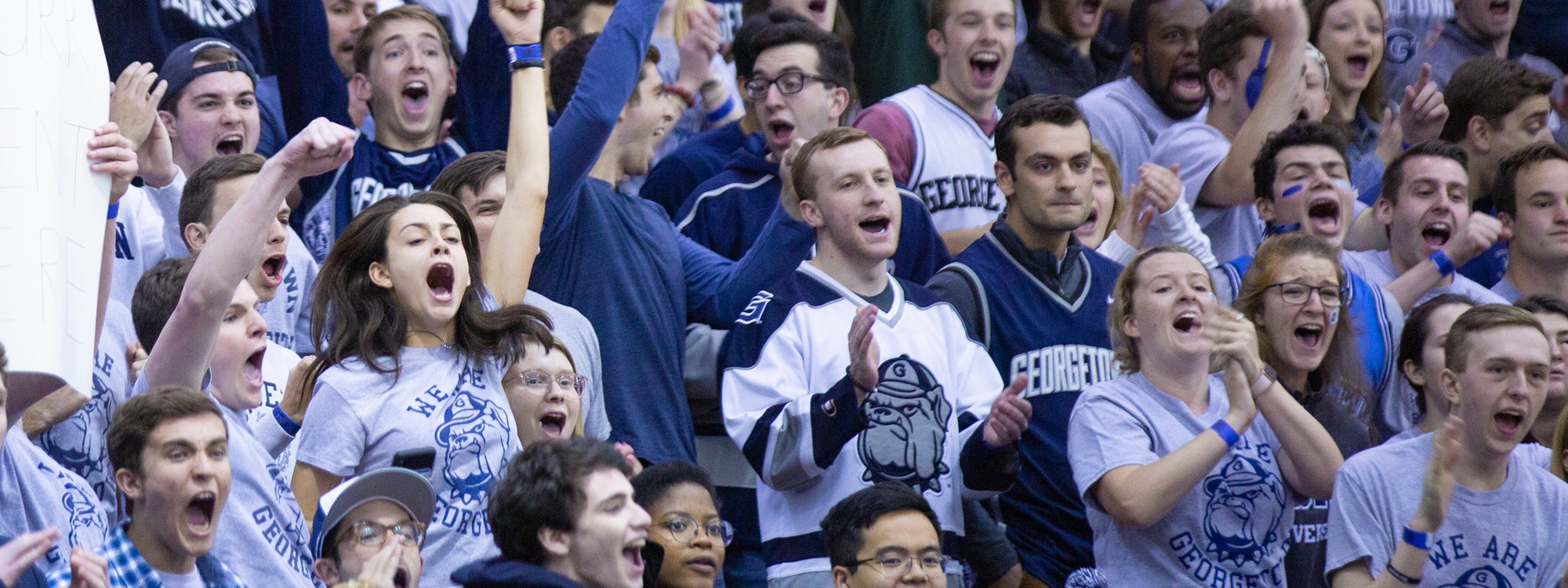 A group of students wearing Georgetown gear cheer enthusiastically at a basketball game.