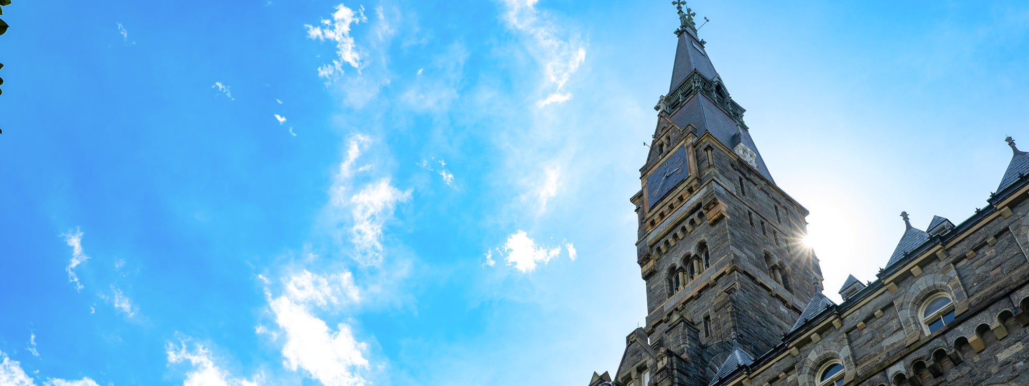 The Healy Hall clocktower in front of a bright blue sky with sun shining behind it.