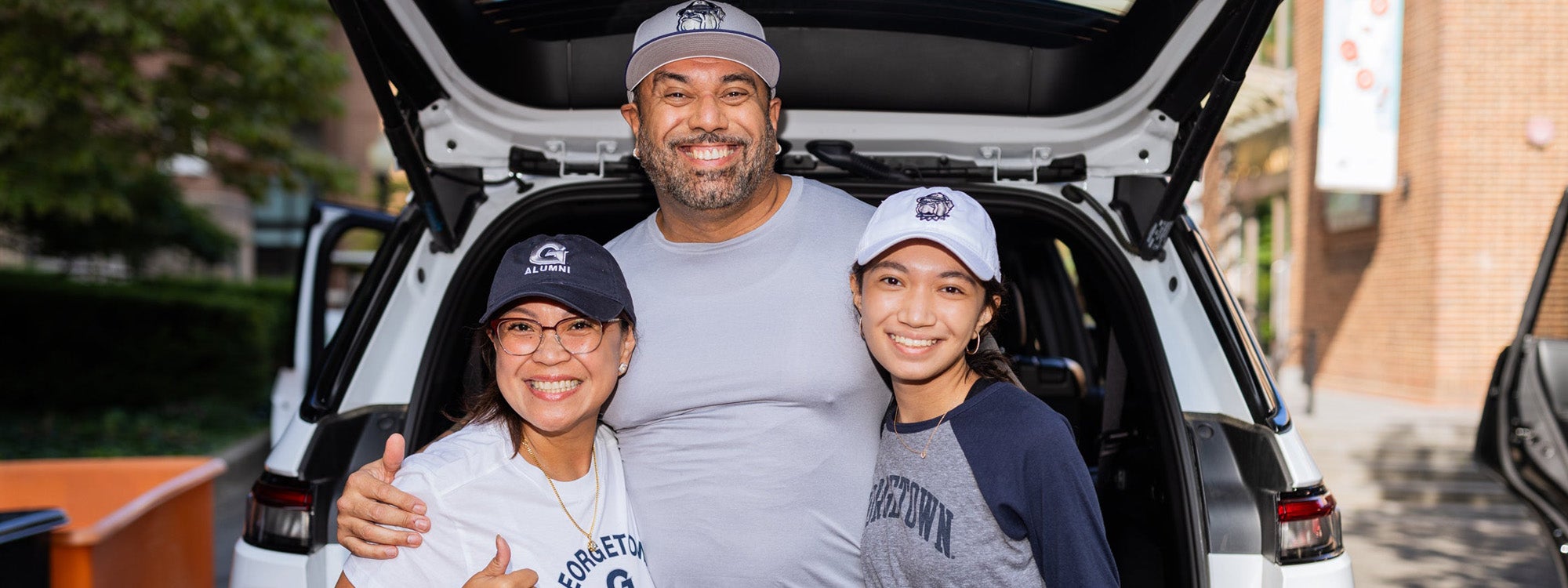 A family embraces in front of an open car trunk during move-in. The family are all wearing Georgetown shirts and hats.